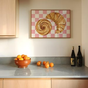 K&ouml;nnte beinhalten: Ein gerahmter Druck eines Croissants und eines Strudelgeb&auml;cks auf einem rosa-wei&szlig;en karierten Hintergrund. Der Druck h&auml;ngt an einer wei&szlig;en Wand &uuml;ber einer K&uuml;chentheke mit einer Sch&uuml;ssel Orangen und zwei Flaschen Wein.