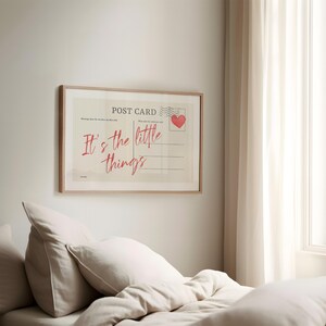 Pu&ograve; includere: Stampa incorniciata di una cartolina con la scritta "It's the little things" in rosso. Il design della cartolina include un timbro a forma di cuore rosso e linee per la scrittura. La cornice &egrave; in legno chiaro, appesa a una parete bianca.