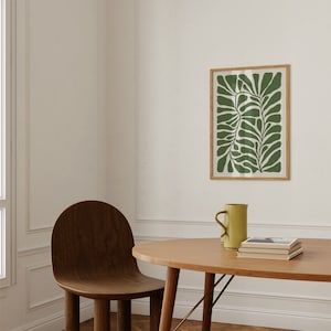 May include: A wooden chair with a rounded back and a light wood table with a green framed print of a stylised leaf design above it. A yellow ceramic mug sits on the table.