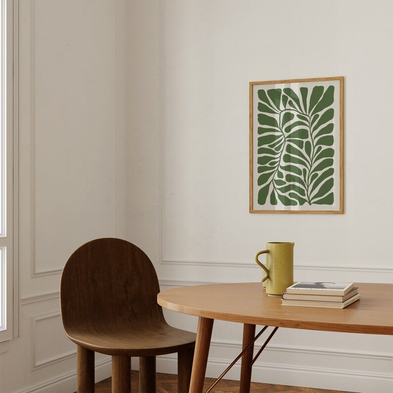 May include: A wooden chair with a rounded back and a light wood table with a green framed print of a stylised leaf design above it. A yellow ceramic mug sits on the table.
