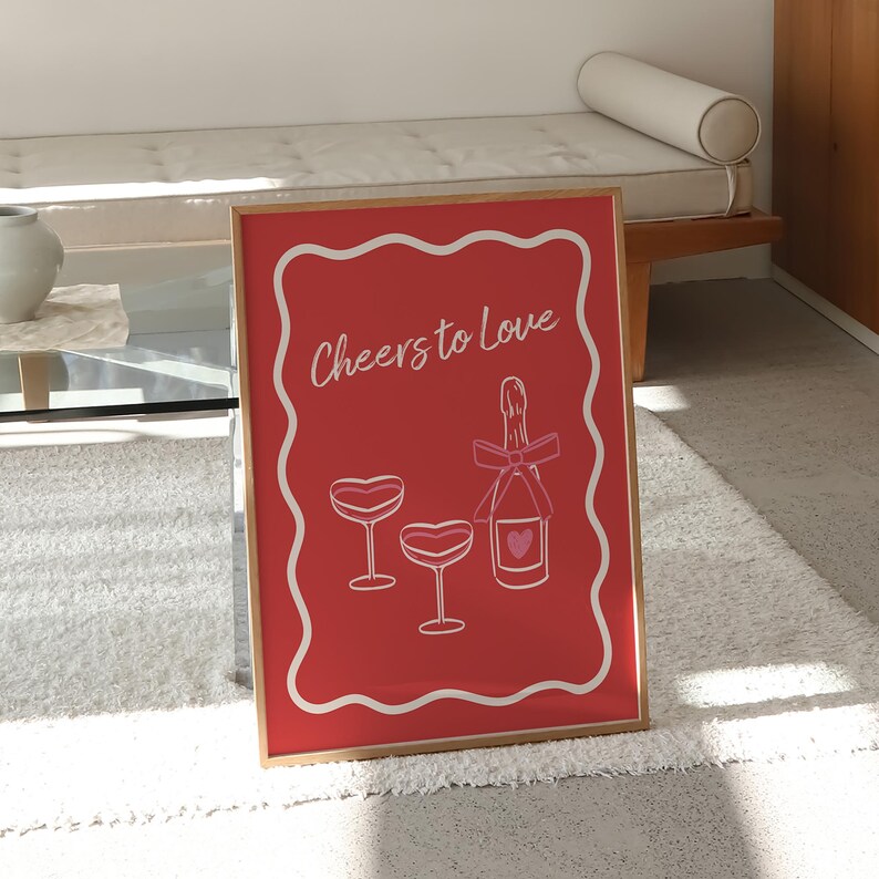 K&ouml;nnte beinhalten: Ein roter gerahmter Druck mit einem wei&szlig;en Rahmen und dem Text "Cheers to Love" in Wei&szlig;. Der Druck zeigt eine Flasche Champagner mit einem rosa Herzen und zwei Champagnergl&auml;ser mit rosa Herzen.