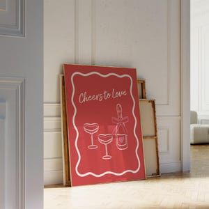 K&ouml;nnte beinhalten: Ein roter Poster mit einem wei&szlig;en Rahmen und dem Text "Cheers to Love" in wei&szlig;. Der Poster zeigt eine wei&szlig;e Illustration einer Champagnerflasche und zwei Champagnergl&auml;ser mit Herzen.