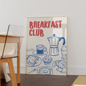 Op de afbeelding: Een ingelijste print met een gouden rand. De print toont een blauw-witte illustratie van ontbijtproducten, waaronder een kopje koffie, toast, een muffin, een croissant en een gebakken ei. De woorden "BREAKFAST CLUB" staan bovenaan in het rood.