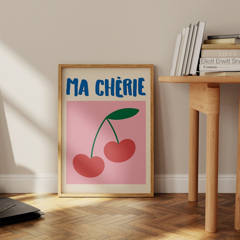 May include: A pink poster with a wooden frame featuring a graphic of two red cherries with green stems and leaves. The poster has the text "MA CHERIE" in blue letters.