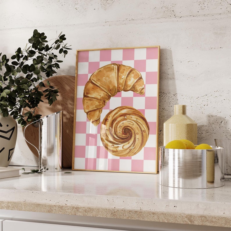 K&ouml;nnte beinhalten: Ein gerahmter Druck mit einem rosa-wei&szlig;en karierten Hintergrund, der eine Aquarell-Illustration eines Croissants und eines Strudels aus Geb&auml;ck zeigt.