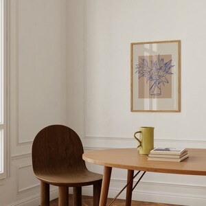 May include: A wooden table with a light brown oval tabletop and a brown wooden chair. A light brown mug and a stack of books are on the table. A framed print of a blue line drawing of flowers in a vase is on the wall behind the table.