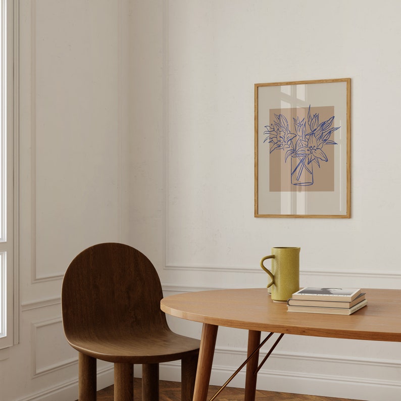 May include: A wooden table with a light brown oval tabletop and a brown wooden chair. A light brown mug and a stack of books are on the table. A framed print of a blue line drawing of flowers in a vase is on the wall behind the table.