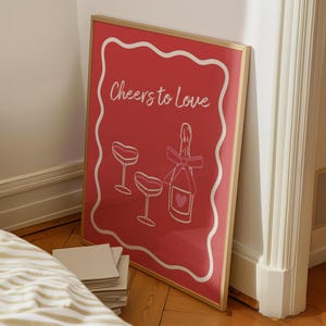 K&ouml;nnte beinhalten: Ein roter gerahmter Druck mit einem wei&szlig;en Rahmen und dem Text "Cheers to Love" in wei&szlig;er Schrift. Der Druck zeigt eine rosafarbene Champagnerflasche mit einem Herzen und zwei rosafarbene Champagnergl&auml;ser.