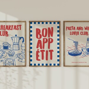 Op de afbeelding: Drie ingelijste kunstprints met een retro-ontwerp. De prints tonen rode en blauwe illustraties en tekst op een cr&egrave;mekleurige achtergrond. E&eacute;n zegt "Breakfast Club", een andere "Bon App&eacute;tit" en de derde "Pasta and Wine Lover Club". De frames zijn lichtbruin.
