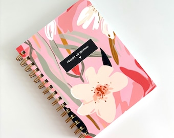 May include: A spiral-bound journal with a pink cover featuring a floral pattern in shades of white, red, and green. A black rectangular label on the front displays the white text "HEALTHY ME JOURNAL" with a small sprout icon underneath.