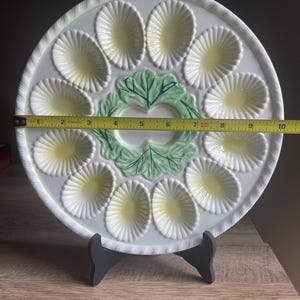 May include: A white ceramic egg serving platter with twelve oval indentations featuring pale yellow centers and a central green leaf design. A yellow measuring tape placed across the center shows the platter has a diameter of 10 inches.