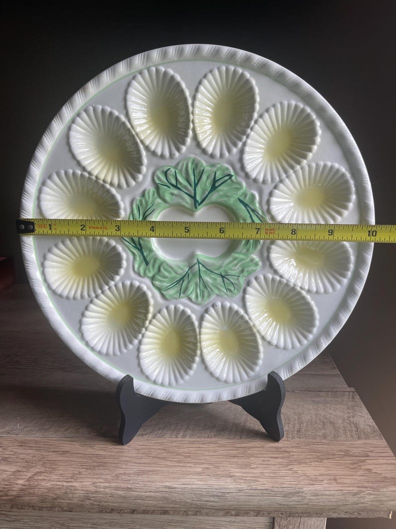 May include: A white ceramic egg serving platter with twelve oval indentations featuring pale yellow centers and a central green leaf design. A yellow measuring tape placed across the center shows the platter has a diameter of 10 inches.