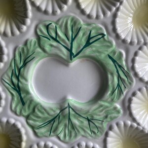 May include: A ceramic oyster plate featuring a central pale green leaf motif with dark green painted veins. The plate is surrounded by multiple scalloped, shell-shaped indentations in a creamy white finish with soft yellow centers for serving.