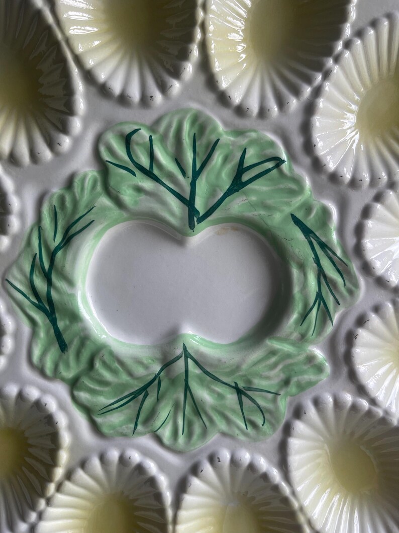 May include: A ceramic oyster plate featuring a central pale green leaf motif with dark green painted veins. The plate is surrounded by multiple scalloped, shell-shaped indentations in a creamy white finish with soft yellow centers for serving.