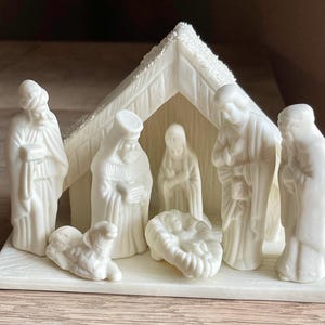 May include: A white nativity scene figurine set featuring a stable with a textured roof and several figures including a central infant in a manger, a lamb, and standing figures arranged on a rectangular base, all crafted in a solid matte white finish.