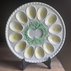 May include: A round white ceramic egg serving platter featuring twelve oval-shaped indentations with pale yellow centers. The center of the plate is decorated with a raised green lettuce leaf design with dark green veining and a scalloped edge.
