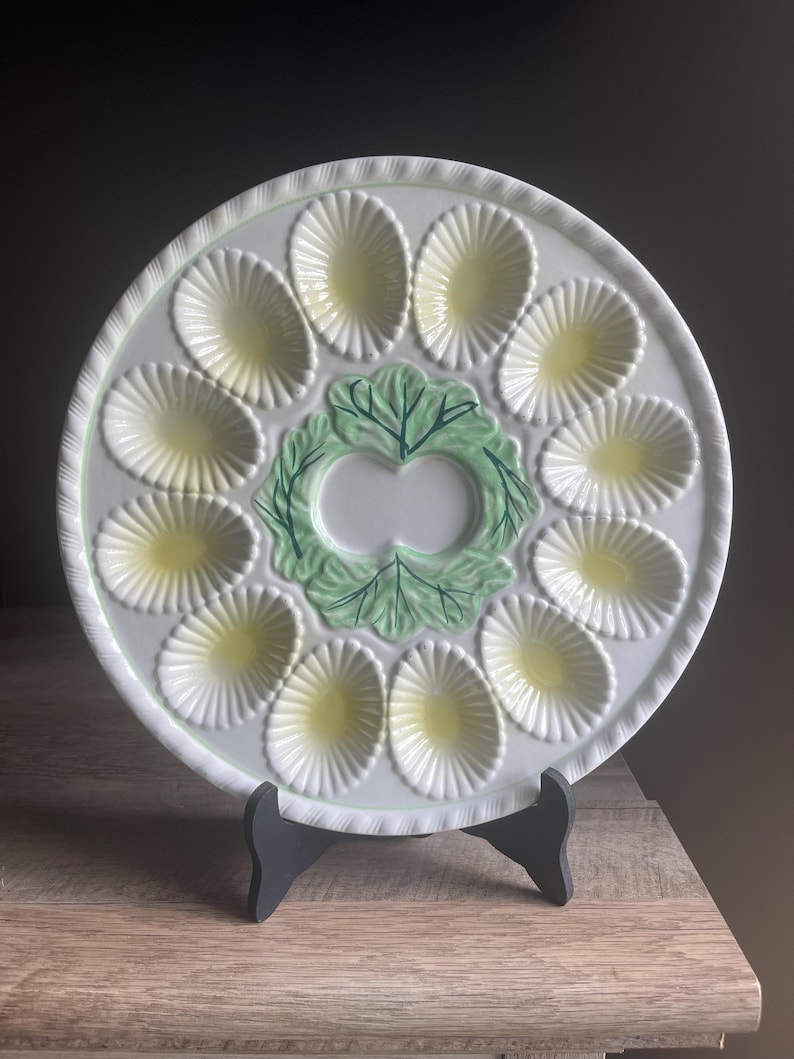 May include: A round white ceramic egg serving platter featuring twelve oval-shaped indentations with pale yellow centers. The center of the plate is decorated with a raised green lettuce leaf design with dark green veining and a scalloped edge.