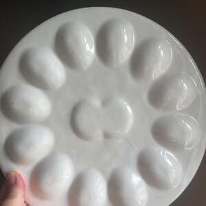 May include: A round, white ceramic egg serving platter featuring twelve oval-shaped indentations arranged in a circular pattern around a central, double-lobed depression. The surface has a smooth, glossy finish with a clean, minimalist aesthetic.