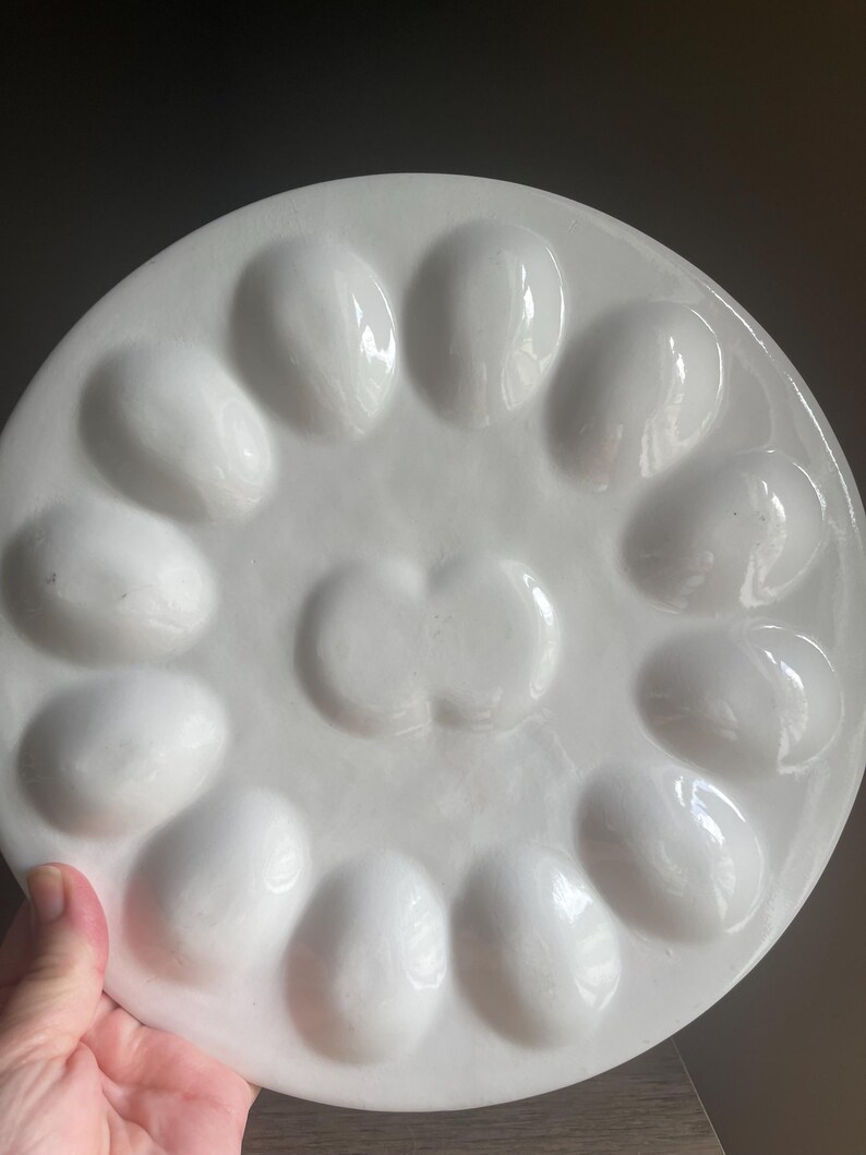 May include: A round, white ceramic egg serving platter featuring twelve oval-shaped indentations arranged in a circular pattern around a central, double-lobed depression. The surface has a smooth, glossy finish with a clean, minimalist aesthetic.