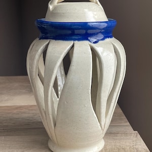 May include: A ceramic lantern with a cream-colored body and a blue band around the top. The lantern has a lid with a twisted handle. The body features vertical cut-out designs. The lantern is sitting on a wooden surface.