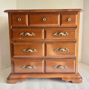 May include: A wooden jewelry chest with a medium brown finish featuring seven drawers. The top row has three small drawers with round knobs, while the three lower rows each contain two wider drawers with ornate, curved brass-toned metal handles.