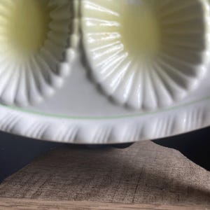 May include: A white ceramic egg tray featuring multiple oval-shaped indentations with a sunburst pattern. The edges of the tray are decorated with a delicate, repeating scalloped texture and a thin, pale green line encircling the perimeter.