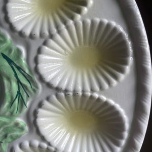 May include: A white ceramic oyster plate featuring multiple oval-shaped, fluted indentations for serving. The plate includes a decorative relief element on the side painted in light green with dark green accents, resembling foliage or seaweed.