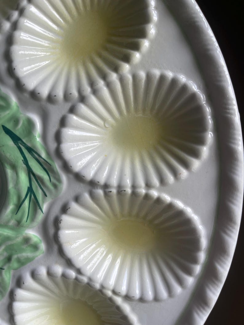 May include: A white ceramic oyster plate featuring multiple oval-shaped, fluted indentations for serving. The plate includes a decorative relief element on the side painted in light green with dark green accents, resembling foliage or seaweed.