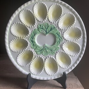 May include: A round white ceramic oyster plate featuring twelve shell-shaped indentations with pale yellow centers arranged in a circle around a central green embossed lettuce leaf design. The plate has a scalloped edge and a textured rim.