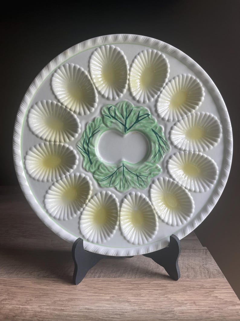 May include: A round white ceramic oyster plate featuring twelve shell-shaped indentations with pale yellow centers arranged in a circle around a central green embossed lettuce leaf design. The plate has a scalloped edge and a textured rim.