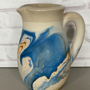 May include: A ceramic pitcher with a spout and handle. The pitcher features a design of blue, white, and tan swirls. The top and handle are a solid tan color. The pitcher is sitting on a gray surface.