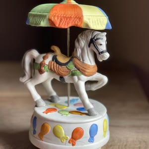 May include: A ceramic carousel music box featuring a white horse with a black bridle and saddle. The horse is under a colorful umbrella. The base is decorated with balloons in various colors. A charming decorative piece.