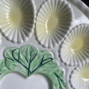 May include: A white ceramic serving platter featuring multiple oval-shaped indentations with pale yellow centers. The platter is decorated with a raised, light green cabbage leaf design that includes dark green painted veins and details.