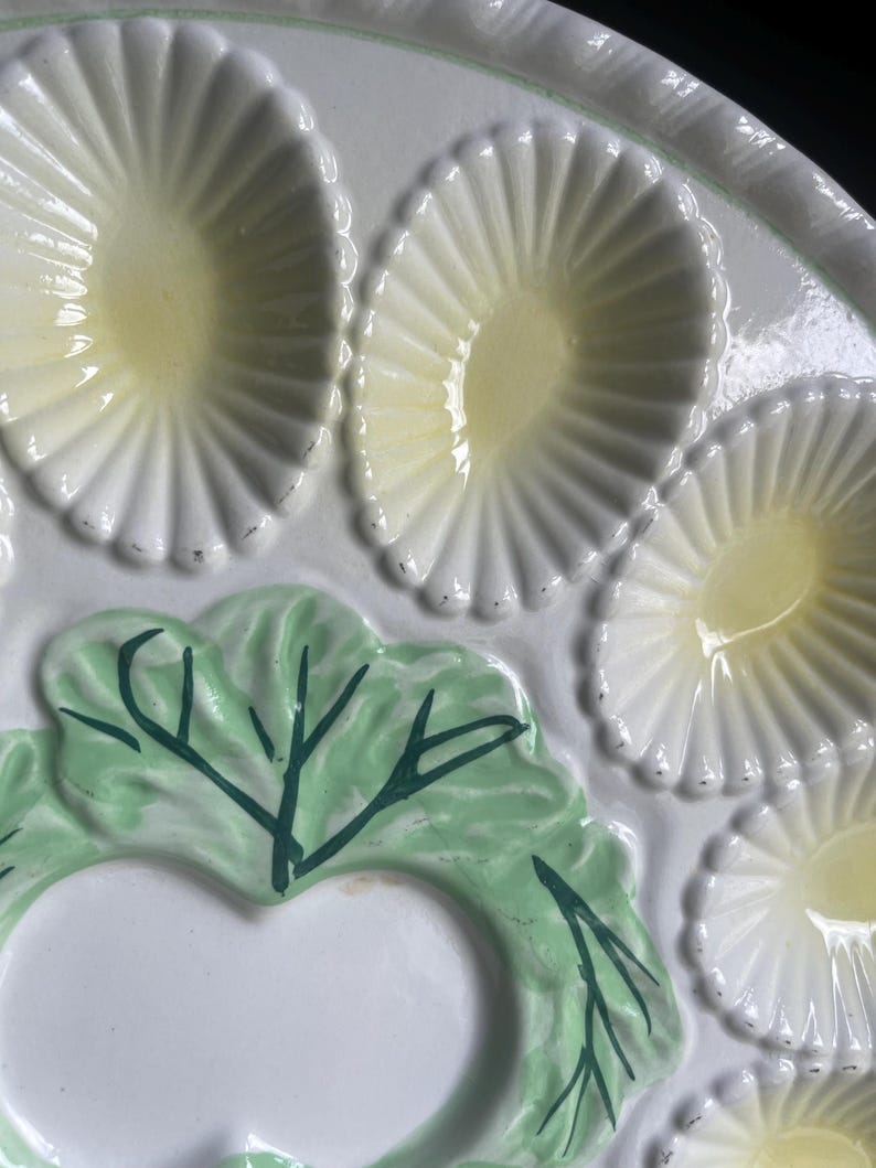 May include: A white ceramic serving platter featuring multiple oval-shaped indentations with pale yellow centers. The platter is decorated with a raised, light green cabbage leaf design that includes dark green painted veins and details.