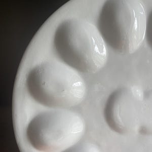 May include: A white ceramic egg serving platter featuring a circular arrangement of twelve smooth, oval-shaped indentations designed to hold individual eggs. The surface has a glossy, reflective finish with a clean, minimalist aesthetic.