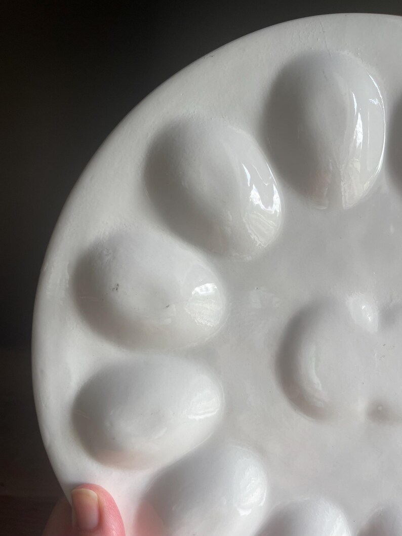 May include: A white ceramic egg serving platter featuring a circular arrangement of twelve smooth, oval-shaped indentations designed to hold individual eggs. The surface has a glossy, reflective finish with a clean, minimalist aesthetic.