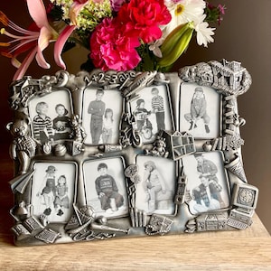 May include: A silver-colored multi-photo frame with eight rectangular openings, each holding a black and white photograph. The frame is decorated with various embossed objects, including toys and school-themed items. A bouquet of pink and red flowers sits behind the frame.