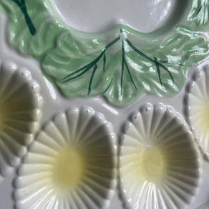May include: A white ceramic oyster plate featuring a glossy finish with molded shell-shaped indentations. Each indentation has a pale yellow center and white radiating ridges. The plate is decorated with raised green leaf accents.