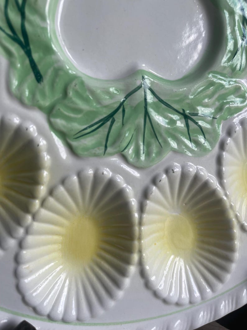 May include: A white ceramic oyster plate featuring a glossy finish with molded shell-shaped indentations. Each indentation has a pale yellow center and white radiating ridges. The plate is decorated with raised green leaf accents.