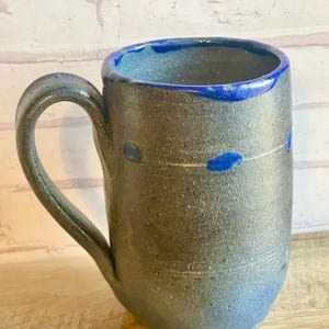 May include: A handmade ceramic mug with a dark gray exterior and a blue interior and rim. The mug features a curved handle and is adorned with blue accents. The bottom of the mug fades to a lighter blue. The mug is approximately 5 inches tall.