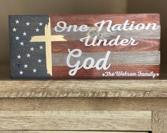 May include: Wooden sign with a distressed American flag design. The sign reads "One Nation Under God" with a gold cross and the text "The Watson Family" at the bottom.