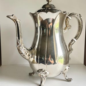 May include: A polished silver-toned metal teapot featuring a curved spout with ornate detailing, a matching handle, and a lid topped with a decorative finial. The body of the teapot has a smooth, reflective surface and stands on three feet.