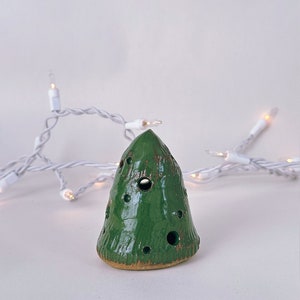 May include: A small green ceramic Christmas tree with holes throughout. The tree is sitting on a white surface with white Christmas lights in the background.