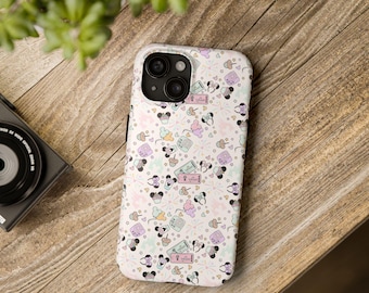 May include: A white phone case with a repeating pattern of cartoon characters, cupcakes, hearts, and other small icons in pastel colors. The case is on a wooden surface, with a camera and plant visible in the background.