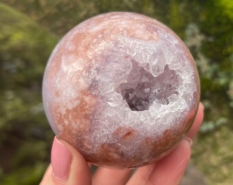 Puede incluir: Una esfera de ágata rosa y marrón con una gran cavidad de geoda abierta. La geoda está revestida de cristales blancos.