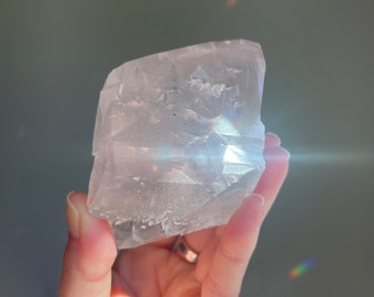 May include: A hand holds a large, translucent, light pink crystal. The crystal has a rough, textured surface with visible facets and internal structures. The background is a soft gray, with a few rainbow light refractions.