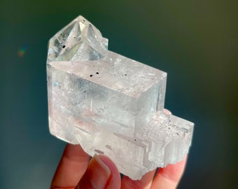 May include: A clear, crystalline mineral specimen with a geometric, layered structure. The translucent stone has a faceted, angular form, with a slightly rough, textured surface. The crystal is held against a blurred, dark green background.