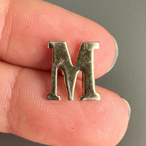 Może przedstawiać: Wyświetlana jest mała, złota litera "M". Wielka litera jest w kroju szeryfowym i wydaje się być urokiem lub wisiorkiem. Tło jest w jednolitym szarym kolorze.