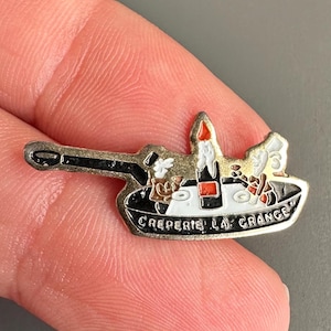 May include: A silver-colored pin in the shape of a tank, featuring a red and white design with the text "Creperie La Grange". The pin has a black cannon and a small, detailed design on top.