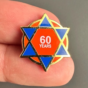 Puede incluir: Un pin dorado con un diseño de estrella de David azul y detalles en naranja. El centro de la estrella muestra el número "60" y la palabra "YEARS" en texto blanco. El pin está sobre un fondo gris.